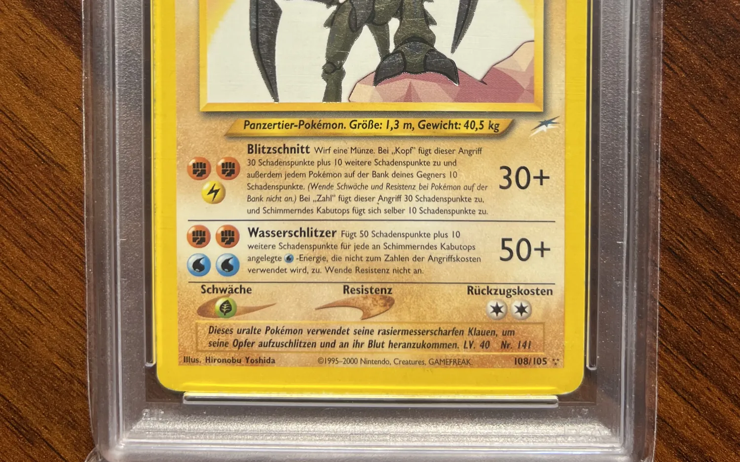 Unterer Teil einer deutschen Pokémon-Karte, Schimmerndes Kabutops, im Slab: Attacken, Unterkante und beide Ecken der Karte im Gehäuse auf einem Holztisch.