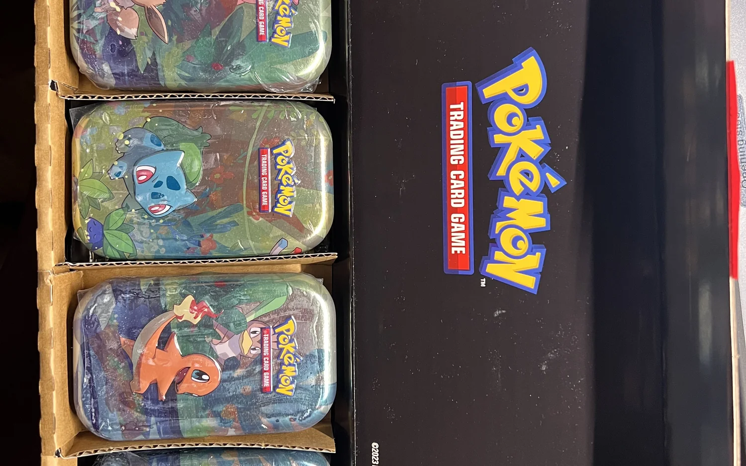 Ein geöffneter Karton mit eingeschweißten Pokémon-Minidosen neben einem schwarzen Display, wie ein Händler seine Ware zu einer Kartenmesse mitbringt.
