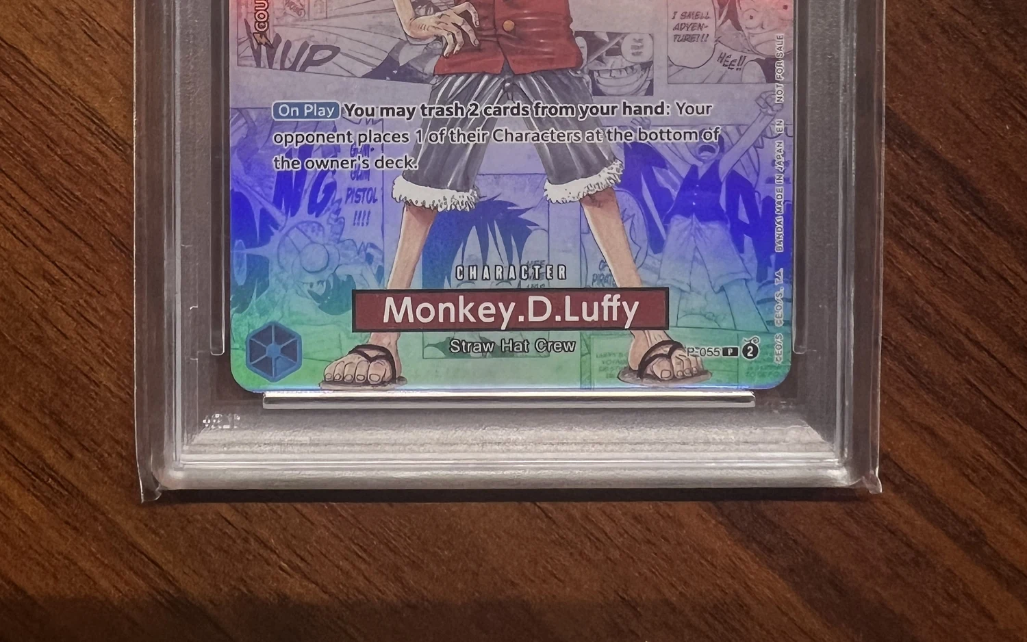 Unterer Teil einer One-Piece-Karte, Monkey D. Luffy, im Slab: Namensleiste und Unterkante der Karte im Gehäuse auf einem Holztisch.