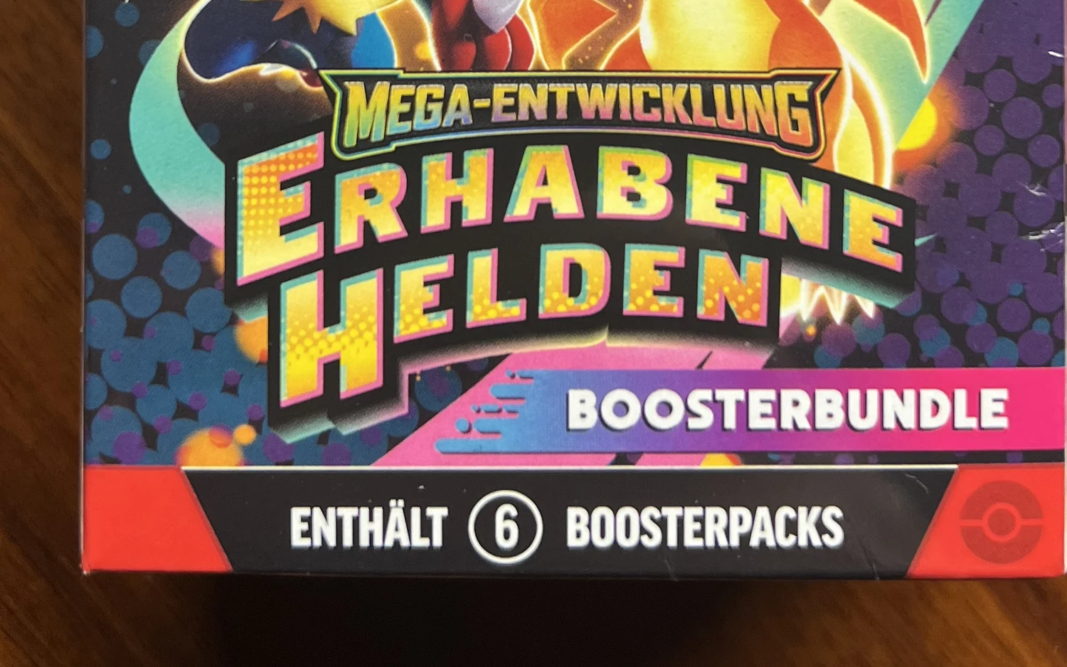 Unterer Teil eines versiegelten Pokémon-Boosterbundles Mega-Entwicklung Erhabene Helden mit dem Aufdruck Enthält 6 Boosterpacks auf einem Holztisch.