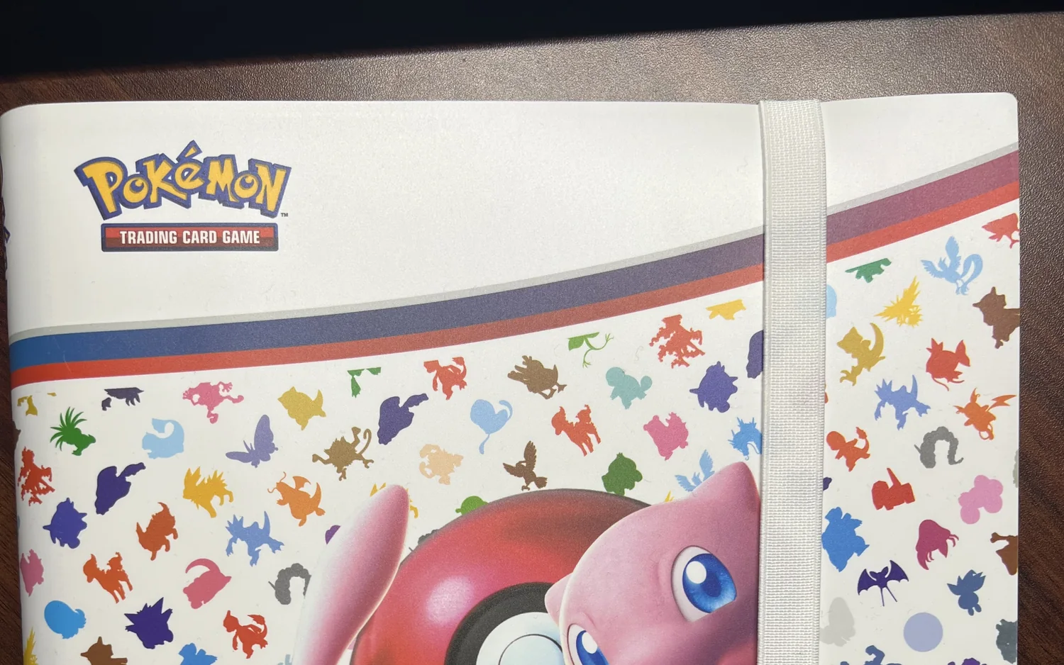 Ein weißes Pokémon-Sammelalbum auf einem Holztisch, oben das Logo des Sammelkartenspiels, darunter bunte Pokémon-Silhouetten und ein Gummiband als Verschluss.