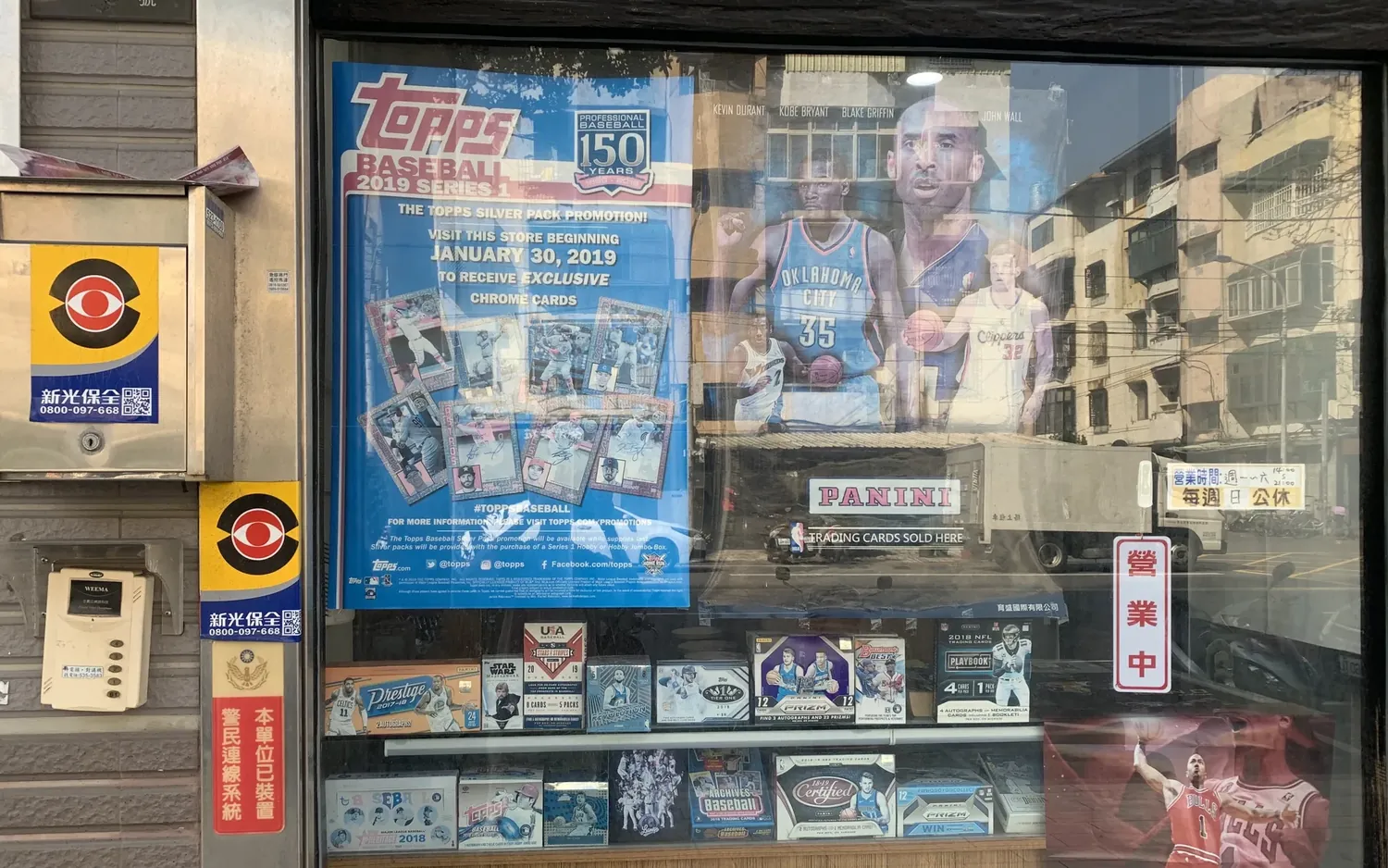 Das Schaufenster eines Kartenladens mit Werbeplakaten und ausgelegten Sammelkartenpackungen von Topps und Panini.