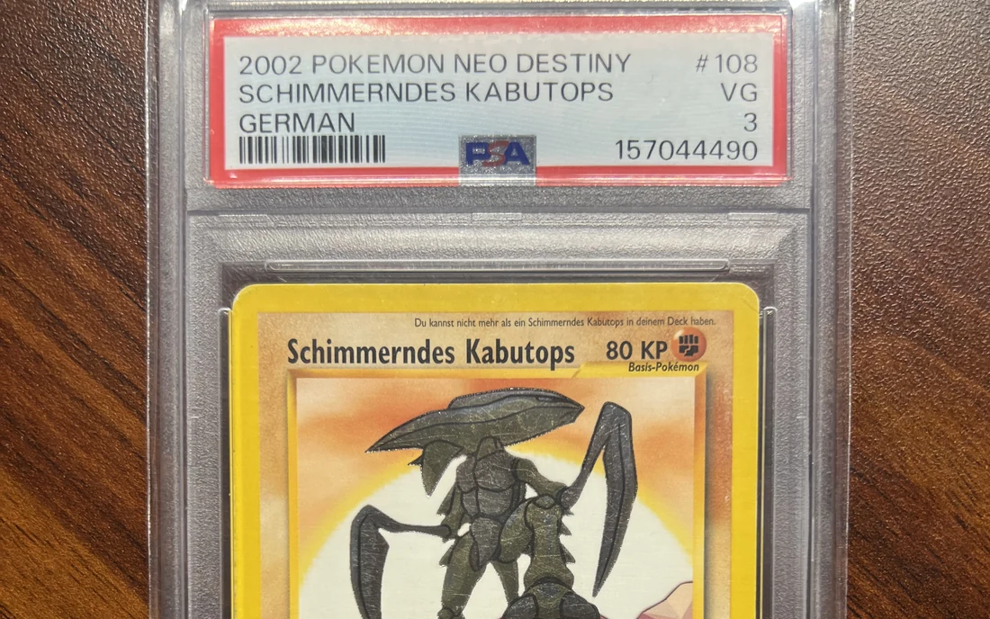 Eine gegradete Pokémon-Karte im PSA-Gehäuse auf einem Holztisch, das Etikett nennt Schimmerndes Kabutops aus Neo Destiny von 2002 in der Note VG 3.