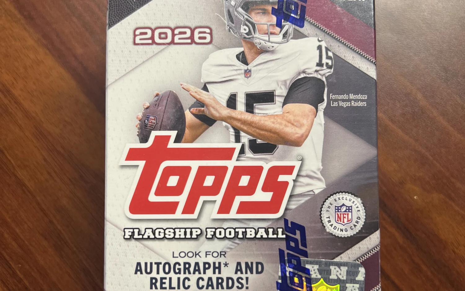 Versiegelte Box Topps Flagship Football 2026 auf einem Holztisch, mit Quarterback der Las Vegas Raiders und dem Hinweis auf Autogramm- und Relic-Karten