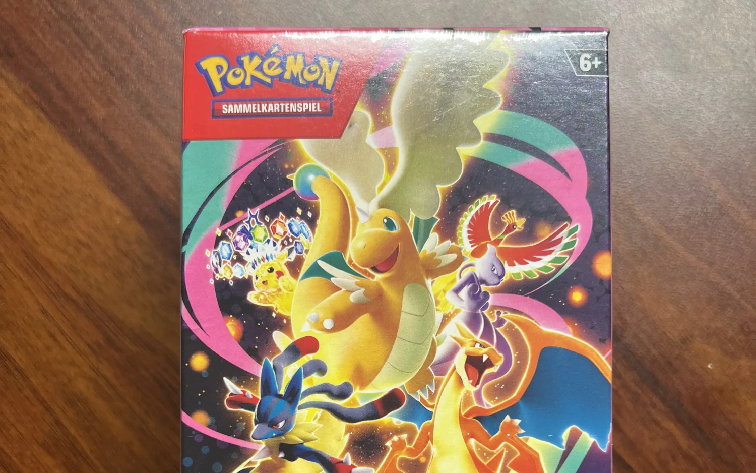 Ein verschlossener Karton des Pokémon-Sammelkartenspiels auf einem Holztisch, auf der Vorderseite eine Illustration mit mehreren Pokémon und die Altersangabe ab sechs Jahren.